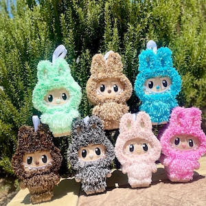 May include: A selection of plush bunny-eared figures in shades of green, brown, blue, pink, and grey. Each figure has a white face with large eyes and a small mouth. They are designed to be ornaments.