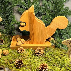 Op de afbeelding: Houten eekhoorn notenkraker met een bruine afwerking. De eekhoorn houdt een noot in zijn bek en heeft een staart die om zijn lichaam gekruld is. De notenkraker staat op een houten basis met een kleine stapel noten ervoor.