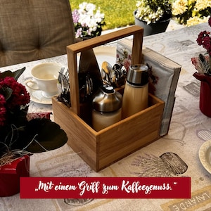 Op de afbeelding: Een rechthoekige houten mand met een centraal handvat, gevuld met twee glazen dispensers met zilverkleurige deksels, een verzameling zilveren vorken en een stapel servetten. De tekst "Mit einem Griff zum Kaffeegenuss." is zichtbaar.