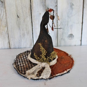 May include: A decorative witch hat crafted from dark brown fabric with a pointed crown. The brim features a mix of black and white checkered fabric and solid orange material. It is adorned with a white fabric tie, dried greenery, and a small bell.