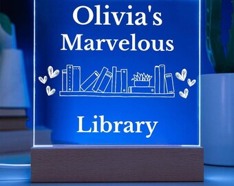 May include: A blue LED light with a white wooden base. The light features a white inscription that reads "Olivia's Marvelous Library" with a drawing of a bookshelf and books.