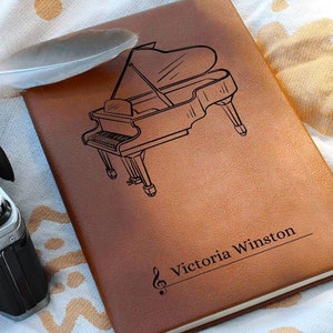 May include: A brown leather journal with a black engraved image of a grand piano and the text "Victoria Winston" with a treble clef symbol.