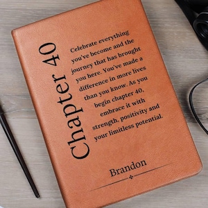May include: A brown leather journal with the text "Chapter 40" and inspirational text. The journal includes the name "Brandon" and a ribbon bookmark. A pen and binoculars are also visible.