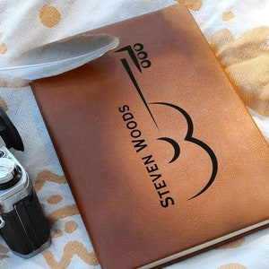 May include: A brown leather journal with the text "STEVEN WOODS" and a guitar graphic engraved on the front cover. The journal is lying on a white and brown patterned blanket.
