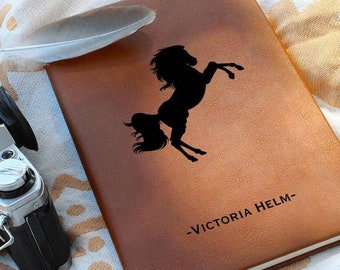 May include: A brown leather journal with a black silhouette of a horse and the text "VICTORIA HELM-." The journal is lying on a white and beige patterned blanket.