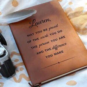 Puede incluir: Un diario de cuero marrón con el texto "Lauren, MAY YOU BE proud OF THE work YOU DO THE person YOU ARE AND THE difference YOU MAKE" grabado en la portada.
