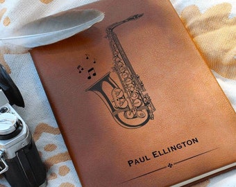 Może przedstawiać: Brązowy skórzany notes z czarną ilustracją saksofonu i nut. Notes ma wygrawerowane imię "PAUL ELLINGTON" na przedniej stronie.