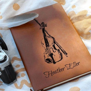 May include: Brown leather journal with a black engraved image of a violin and bow. The journal is personalized with the name "Heather Eller".
