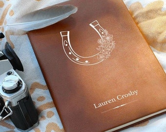 May include: A brown leather journal with a white horseshoe design filled with flowers. The journal has the name "Lauren Crosby" printed on it. A feather and a camera are next to the journal on a patterned blanket.