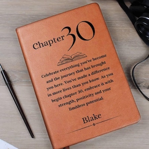 May include: A brown leather journal with the words "Chapter 30" and a personalized name. The journal has a ribbon bookmark and a message about celebrating life's journey. A pen and binoculars are in the background.