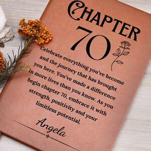 May include: A brown leather journal with the words "CHAPTER 70" in black script. The journal has a floral design and the text: "Celebrate everything you've become..." and the name "Angela". A bookmark and a sprig of orange flowers are visible.