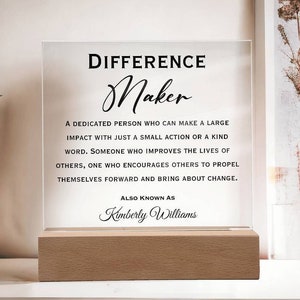 May include: A clear acrylic sign with the text "DIFFERENCE Maker" and a definition of a difference maker. The text also includes "ALSO KNOWN AS Kimberly Williams".