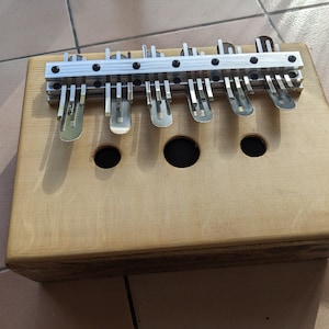 Peut inclure: Un instrument de musique en bois avec une finition naturelle. L'instrument possède une rangée de touches métalliques qui servent à créer des sons. Les touches sont montées sur une barre métallique fixée au corps en bois. L'instrument possède trois trous ronds sur le devant.