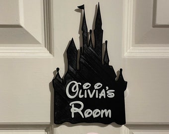 Pode incluir: Placa de porta preta em forma de castelo com o texto "Olivia's Room" em escrita branca. A placa está montada numa porta branca. Um círculo rosa com o texto "Handmade by Fred & Liv Designs" está abaixo.