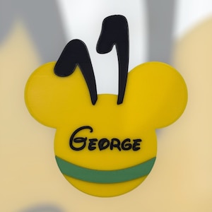 Könnte beinhalten: Gelber Mickey-Maus-förmiger Namensschild mit schwarzen Ohren und dem Namen "George" in schwarzer Kursivschrift. Das Namensschild hat ein grünes Band am unteren Rand.