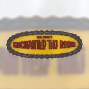 Può includere: Un cartello ovale giallo con un bordo blu e marrone. Il cartello recita "Walt Disney's Enchanted Tiki Room" in lettere rosse.