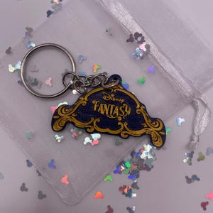 Könnte beinhalten: Ein blauer und goldener Disney Fantasy Schlüsselanhänger mit einem silbernen Schlüsselring. Der Schlüsselanhänger ist in Form eines Schilds mit den Worten "Disney Fantasy" in goldenen Buchstaben.