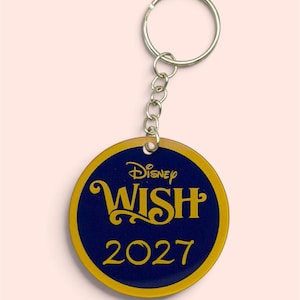 May include: A circular keyring featuring a dark blue centre with a gold border. The text Disney WISH is printed in gold at the top, with the year 2027 printed in gold below it. The keyring is attached to a silver metal split ring by a short chain.
