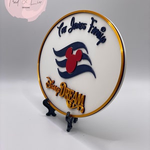 K&ouml;nnte beinhalten: Ein rundes wei&szlig;es Schild mit goldfarbenem Rand und einem goldfarbenen St&auml;nder. Das Schild zeigt ein blaues und wei&szlig;es Mickey Mouse-Kreuzfahrtschiff-Logo mit einem roten Mickey Mouse-Kopf. Der Text "The Jones Family" ist in goldener Kursivschrift &uuml;ber dem Logo geschrieben. Der Text "Disney Dream" ist in goldener Schrift unter dem Logo geschrieben.