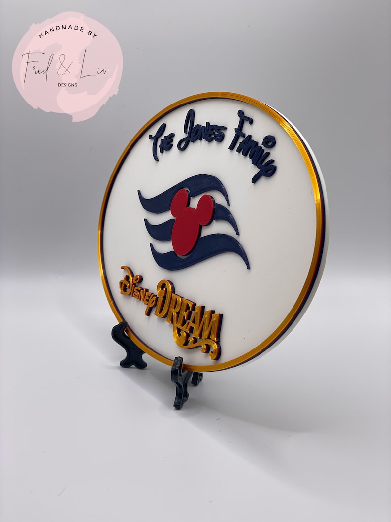 K&ouml;nnte beinhalten: Ein rundes wei&szlig;es Schild mit goldfarbenem Rand und einem goldfarbenen St&auml;nder. Das Schild zeigt ein blaues und wei&szlig;es Mickey Mouse-Kreuzfahrtschiff-Logo mit einem roten Mickey Mouse-Kopf. Der Text "The Jones Family" ist in goldener Kursivschrift &uuml;ber dem Logo geschrieben. Der Text "Disney Dream" ist in goldener Schrift unter dem Logo geschrieben.
