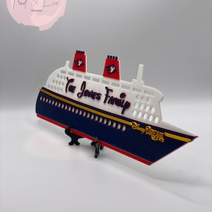 Puede incluir: Un barco decorativo azul marino y blanco con detalles en rojo, con el texto "The Jones Family" y "Disney Dream". El barco tiene dos chimeneas rojas y se exhibe sobre un soporte negro. Las palabras "Handmade by Fred & Liv Designs" est&aacute;n en la esquina superior izquierda.