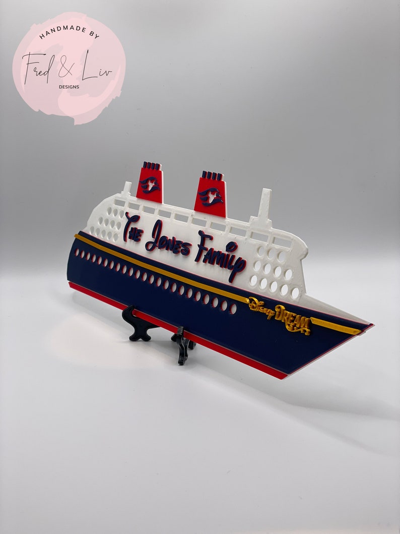 Puede incluir: Un barco decorativo azul marino y blanco con detalles en rojo, con el texto "The Jones Family" y "Disney Dream". El barco tiene dos chimeneas rojas y se exhibe sobre un soporte negro. Las palabras "Handmade by Fred & Liv Designs" est&aacute;n en la esquina superior izquierda.