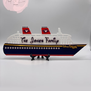 Puede incluir: Un recorte decorativo de un crucero con el texto "The Jones Family" y "Disney Dream". El barco presenta un cuerpo blanco, un casco azul marino, detalles en rojo y adornos dorados. El barco se exhibe sobre un soporte negro.