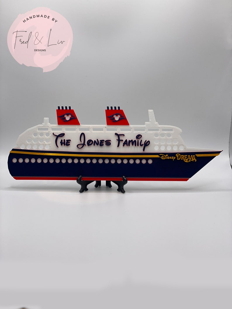 Puede incluir: Un recorte decorativo de un crucero con el texto "The Jones Family" y "Disney Dream". El barco presenta un cuerpo blanco, un casco azul marino, detalles en rojo y adornos dorados. El barco se exhibe sobre un soporte negro.