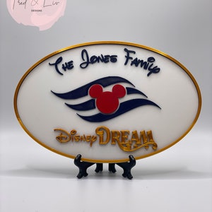 K&ouml;nnte beinhalten: Ein wei&szlig;es ovales Schild mit goldfarbenem Rand. Das Schild zeigt ein blaues und wei&szlig;es Mickey Mouse-Schiffslogo mit einem roten Mickey Mouse-Kopf. Der Text "The Jones Family" ist in Kursivschrift &uuml;ber dem Logo geschrieben. Der Text "Disney Dream" ist in Kursivschrift unter dem Logo geschrieben. Das Schild steht auf einem schwarzen St&auml;nder.