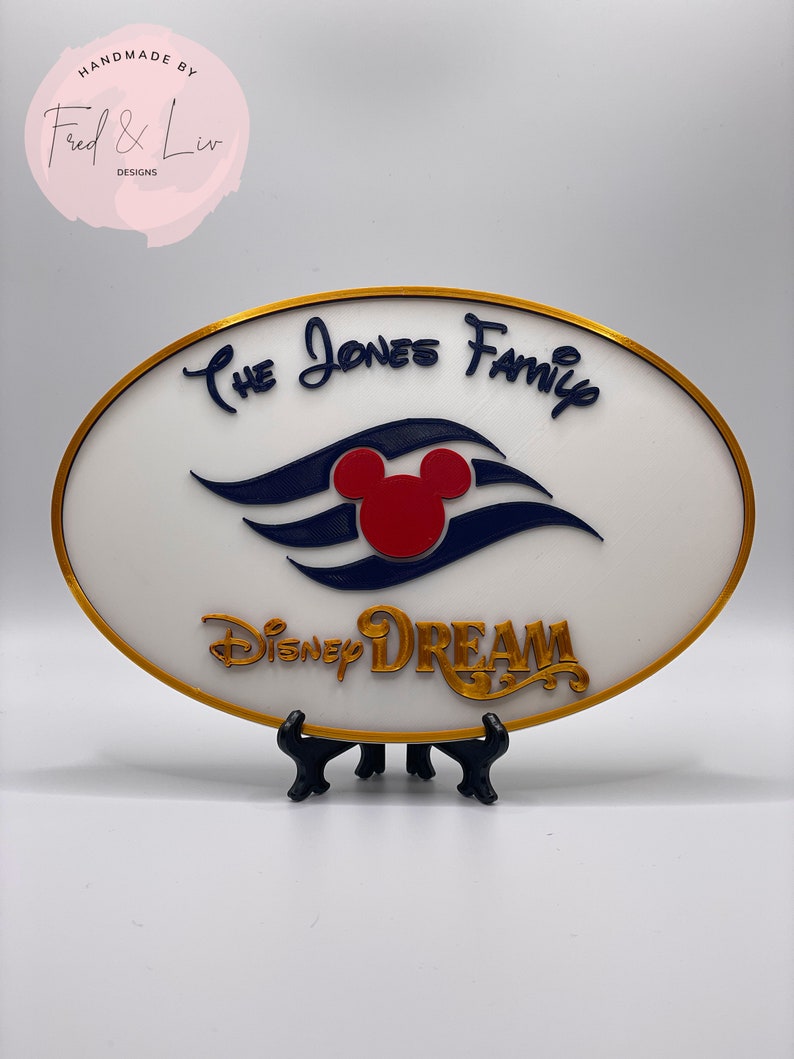 K&ouml;nnte beinhalten: Ein wei&szlig;es ovales Schild mit goldfarbenem Rand. Das Schild zeigt ein blaues und wei&szlig;es Mickey Mouse-Schiffslogo mit einem roten Mickey Mouse-Kopf. Der Text "The Jones Family" ist in Kursivschrift &uuml;ber dem Logo geschrieben. Der Text "Disney Dream" ist in Kursivschrift unter dem Logo geschrieben. Das Schild steht auf einem schwarzen St&auml;nder.