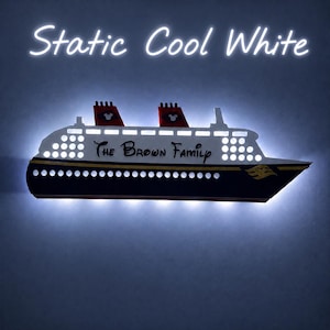 Puede incluir: Decoraci&oacute;n de pared con forma de barco de madera con el texto "The Brown Family" y "Static Cool White". El barco tiene chimeneas rojas y negras con siluetas de Mickey Mouse y est&aacute; iluminado con luces blancas.