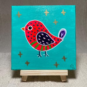 Può includere: Un dipinto su tela quadrata su un piccolo cavalletto di legno raffigurante un uccello rosso con pois neri, un'ala viola scuro con macchie rosa chiaro, strisce verdi e una coda viola. Lo sfondo è turchese con diverse piccole stelle dorate.