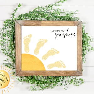 Puede incluir: Una impresión enmarcada con un marco de madera que presenta un sol amarillo y huellas. El texto "you are my sunshine" está escrito en escritura negra. La imagen está rodeada de follaje verde.