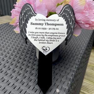 May include: A black heart-shaped memorial plaque with angel wings, a black tie, and pink flowers. The plaque reads "In loving memory of Sammy Thompson 16.12.1939 - 30.09.24 I miss you more than anyone knows. As time goes by the emptiness grows I laugh, I talk, I play, my part. But behind my Smile is a broken Heart."