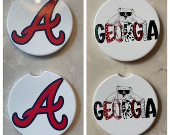 May include: Set of four white round beverage coasters. Two coasters feature a red and blue "A" logo. Two coasters feature a white bulldog wearing sunglasses with the word "GEORGIA" in red, black, and white plaid.
