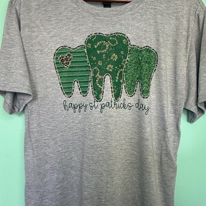 Op de afbeelding: Een grijs T-shirt met een groen, goud en zwart ontwerp van drie tanden. De tanden zijn versierd met klavertjesvier, hoefijzers en harten. De tekst "happy st patricks day" is onder de tanden gedrukt.