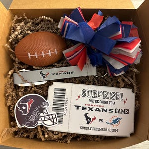 K&ouml;nnte beinhalten: Eine Geschenkbox gef&uuml;llt mit braunem Packpapier, einem rot-wei&szlig;-blauen Pompon, einem Mini-Fu&szlig;ball, einem Schl&uuml;sselanh&auml;nger der Houston Texans und einem Ticket mit der Aufschrift "&Uuml;berraschung! Wir gehen zu einem Spiel der Houston Texans!" mit dem Datum "Sonntag, 15. Dezember 2024".