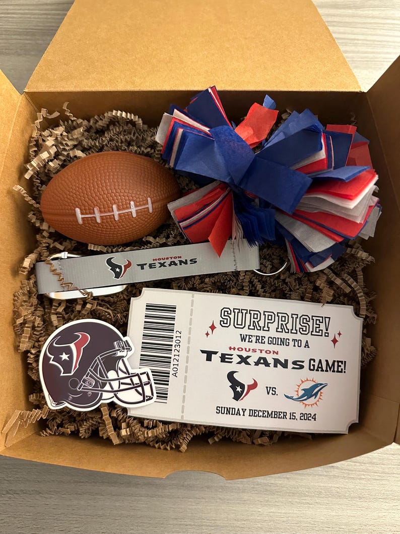 K&ouml;nnte beinhalten: Eine Geschenkbox gef&uuml;llt mit braunem Packpapier, einem rot-wei&szlig;-blauen Pompon, einem Mini-Fu&szlig;ball, einem Schl&uuml;sselanh&auml;nger der Houston Texans und einem Ticket mit der Aufschrift "&Uuml;berraschung! Wir gehen zu einem Spiel der Houston Texans!" mit dem Datum "Sonntag, 15. Dezember 2024".