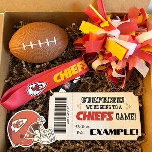 K&ouml;nnte beinhalten: Eine Geschenkbox gef&uuml;llt mit Artikeln zum Thema Kansas City Chiefs, darunter ein roter und gelber Pompon, ein rotes Lanyard mit dem Chiefs-Logo, ein Football, ein Aufkleber eines Chiefs-Helms und ein Ticketstub, auf dem "&Uuml;berraschung! Wir gehen zu einem Chiefs-Spiel!" steht.
