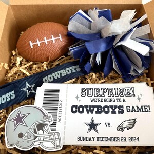 K&ouml;nnte beinhalten: Eine Geschenkbox gef&uuml;llt mit Artikeln zum Thema Dallas Cowboys, darunter ein Football, ein Pompon, ein Lanyard, ein Helm-Aufkleber und ein Ticketstub mit der Aufschrift "&Uuml;berraschung! Wir gehen zu einem Cowboys-Spiel!"  VS.  Sonntag, 29. Dezember 2024.