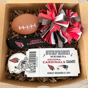 K&ouml;nnte beinhalten: Eine Geschenkbox gef&uuml;llt mit Artikeln zum Thema Arizona Cardinals, darunter ein Football, ein Pompon, ein Stirnband und ein Ticketstub, auf dem steht: "&Uuml;berraschung! Wir gehen zu einem Spiel der Arizona Cardinals!"