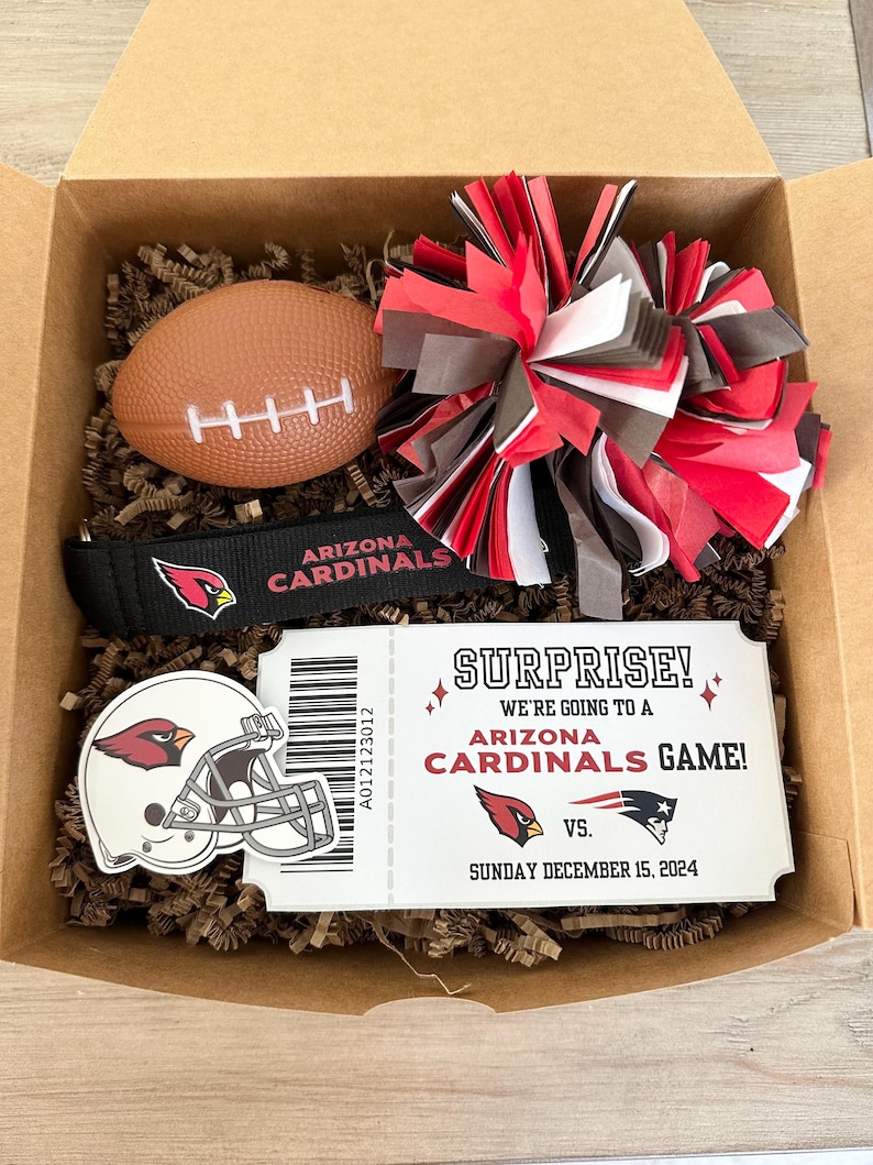 K&ouml;nnte beinhalten: Eine Geschenkbox gef&uuml;llt mit Artikeln zum Thema Arizona Cardinals, darunter ein Football, ein Pompon, ein Stirnband und ein Ticketstub, auf dem steht: "&Uuml;berraschung! Wir gehen zu einem Spiel der Arizona Cardinals!"