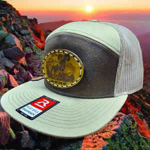 May include: A brown and beige flat bill snapback hat with a brown leather patch featuring a ram and the text "EST 1997 ONE CITY, RAW CUT". The hat has a mesh back panel.