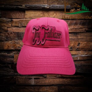 May include: A magenta baseball cap with the text "Tiny Trailblazer" in a brown, layered design. The cap has a curved brim and is set against a wood-grain background. The cap is a solid magenta color.
