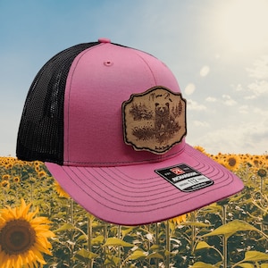 May include: Pink and black trucker hat with a brown leather patch featuring a bear and the text "Mama Bear" in a decorative font. The hat is made by Richardson and has an adjustable strap.