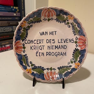 May include: A decorative ceramic plate with a scalloped edge, featuring a floral design in blue, orange, and green. The plate has text in the center that reads "VAN HET CONCERT DES LEVENS KRIJGT NIEMAND EEN PROGRAM".