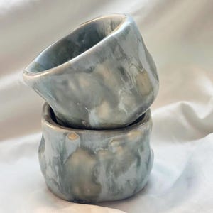 May include: Two ceramic cups stacked vertically, featuring a hand-formed, irregular shape. The glaze is a marbled blend of soft blue, grey, and creamy white tones, creating a smooth, glossy finish across the exterior and interior surfaces.