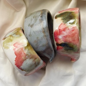 May include: Three ceramic bowls with irregular, organic shapes and glossy finishes. One bowl features a white base with red and green watercolor-style splotches, another is a solid muted grey, and the third has a white base with red and green accents.