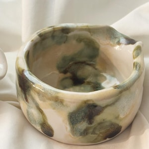 May include: A small, round ceramic bowl with a glossy finish, featuring an irregular, hand-formed rim and a pour spout. The surface displays a marbled glaze pattern in shades of cream, sage green, and dark brown, creating a fluid, organic appearance.