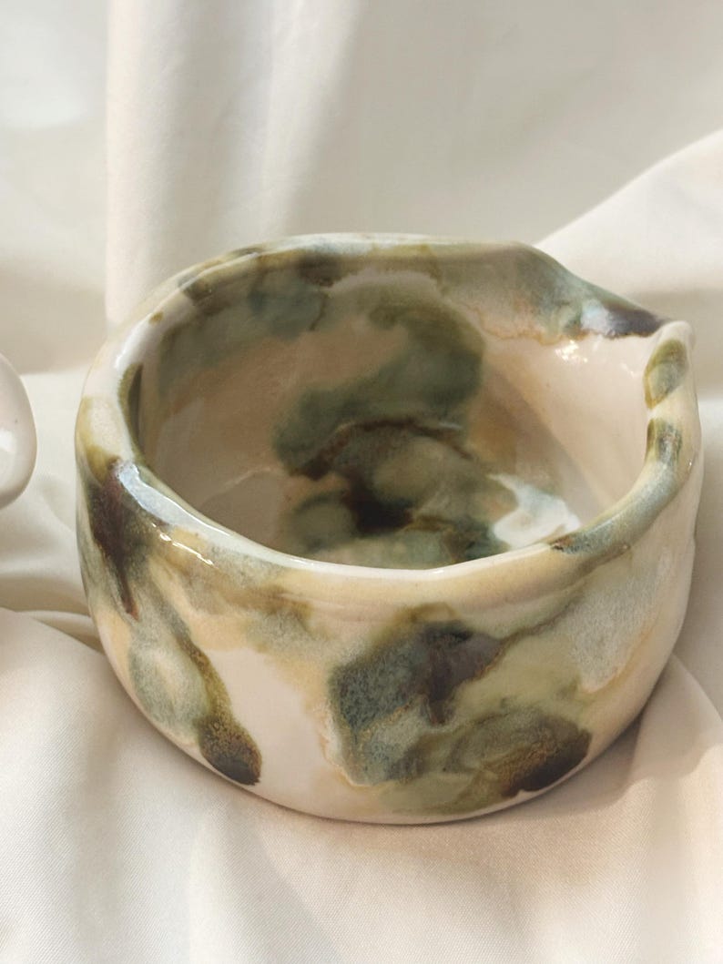 May include: A small, round ceramic bowl with a glossy finish, featuring an irregular, hand-formed rim and a pour spout. The surface displays a marbled glaze pattern in shades of cream, sage green, and dark brown, creating a fluid, organic appearance.